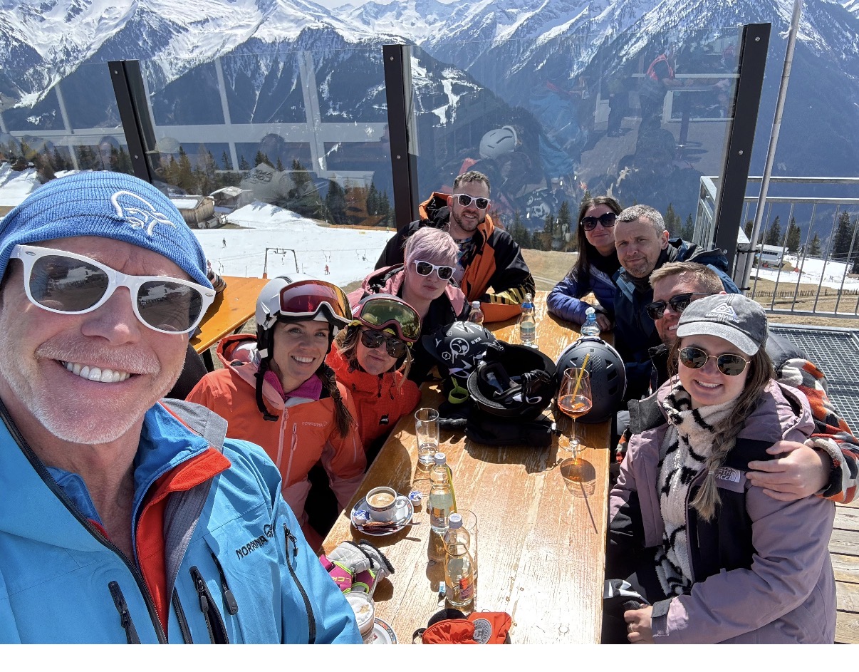 A larger group of EmpoweredFIT friends around a mountain terrace table, with a man in blue taking the selfie
